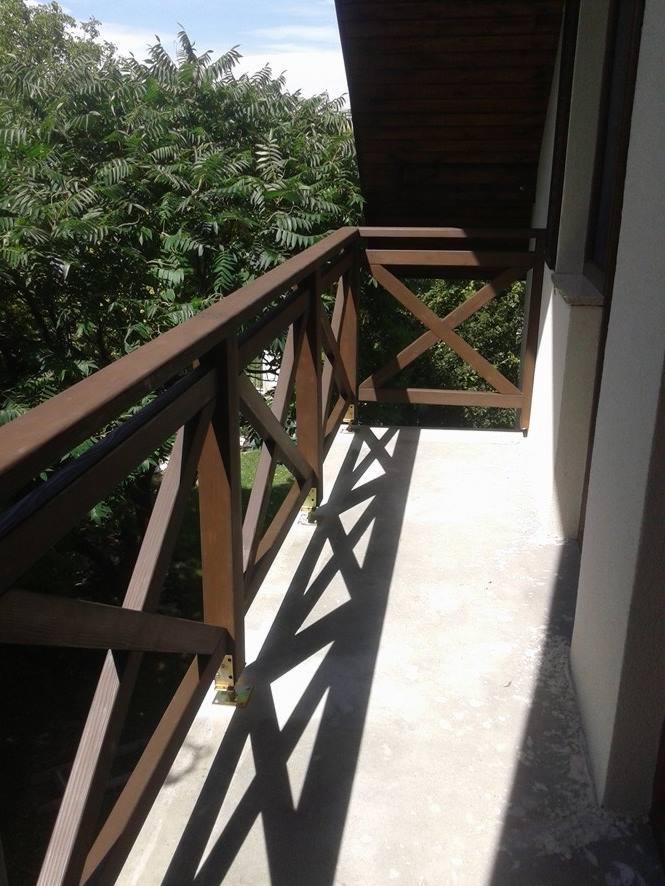 Balkon z drewnianą balustradą o krzyżowym wzorze, pomalowaną na ciemny brąz, rzucającą ostre cienie na betonową podłogę. Widok na zieleń drzew w tle.