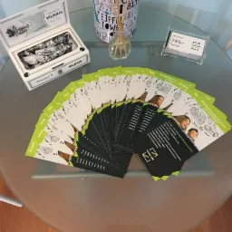 Stos ulotek reklamowych Finance24 na szklanym stoliku, obok puszka cukierków Pergalė Vilnius, dyfuzor zapachowy i wizytownik z wizytówką.