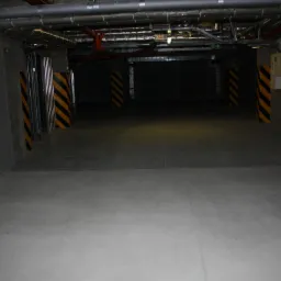 Gładka, szara posadzka w hali garażowej z kolumnami oznaczonymi czarno-żółtymi pasami ostrzegawczymi i widocznymi instalacjami pod sufitem.