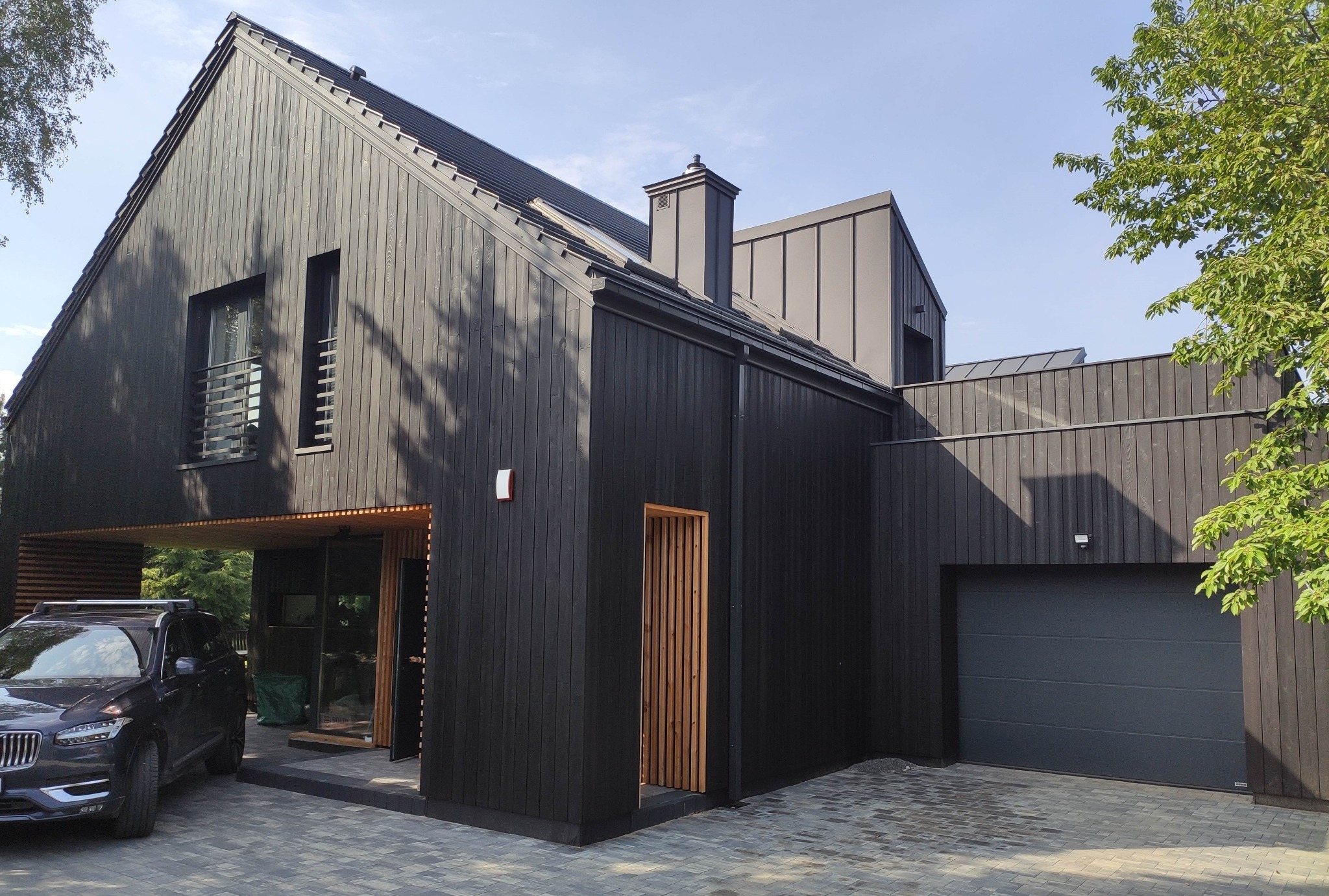 Nowoczesny dom jednorodzinny z czarną drewnianą elewacją, szarym dachem i garażem, z podjazdem wyłożonym kostką brukową i zaparkowanym czarnym SUV-em.