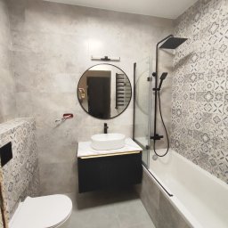 REMONTUJEMY LUBELSKIE Usługi remontowe-wykończenia wnętrz pod klucz - Nowoczesna łazienka z szarymi płytkami imitującymi beton, okrągłym lustrem z czarną ramą, białą umywalką nablatową na czarnej szafce, baterią w kolorze czarnym, oraz wanną z parawanem.