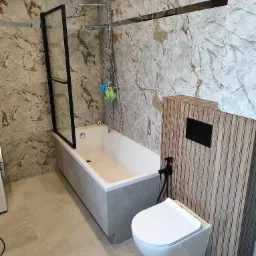 Nowoczesna łazienka z wanną z czarną ramą prysznicową, podwieszaną toaletą z czarną armaturą i płytkami imitującymi marmur na ścianach.