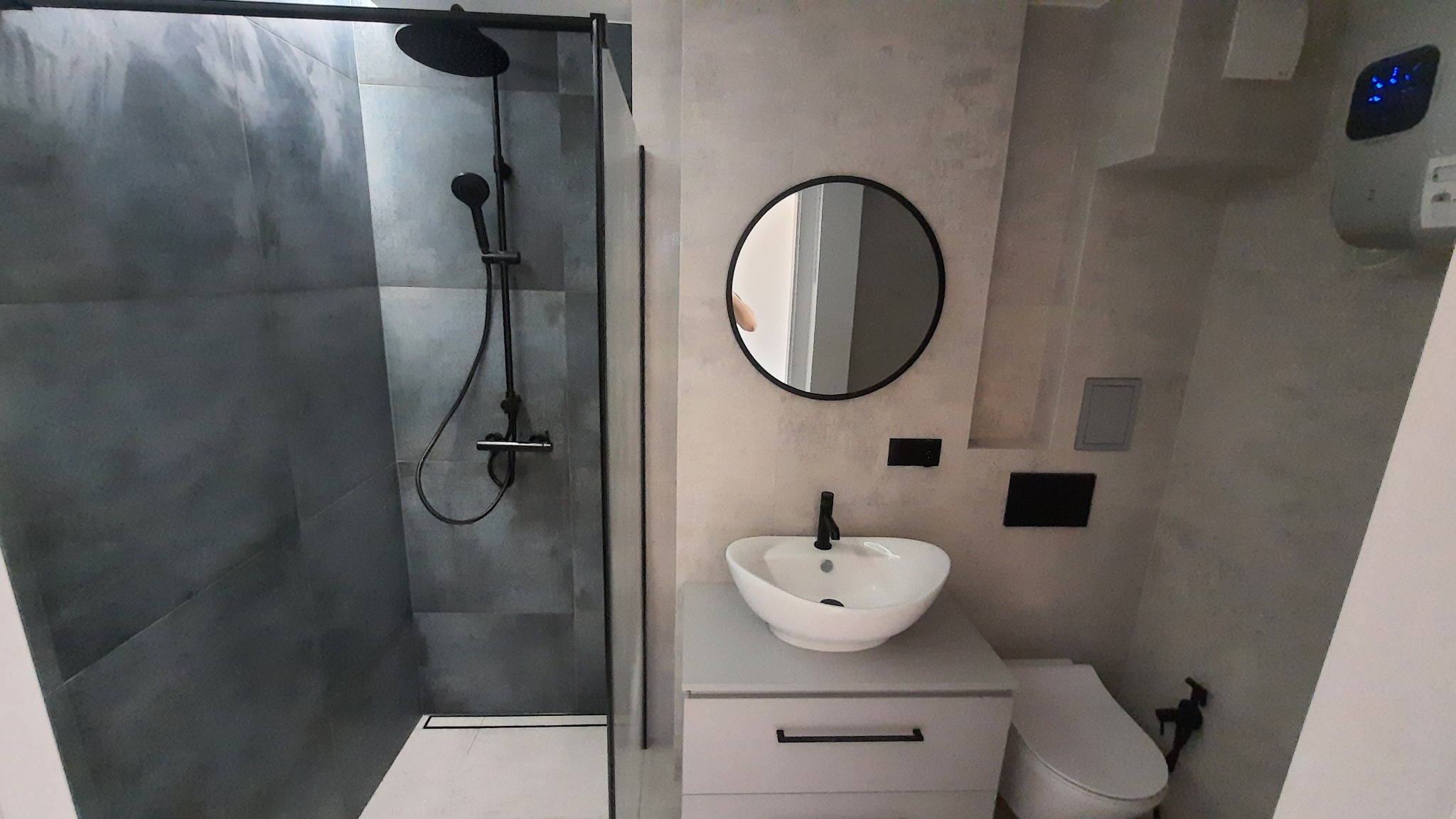 Nowoczesna łazienka z kabiną prysznicową typu walk-in z czarną armaturą, okrągłym lustrem i białą umywalką nablatową na szafce.