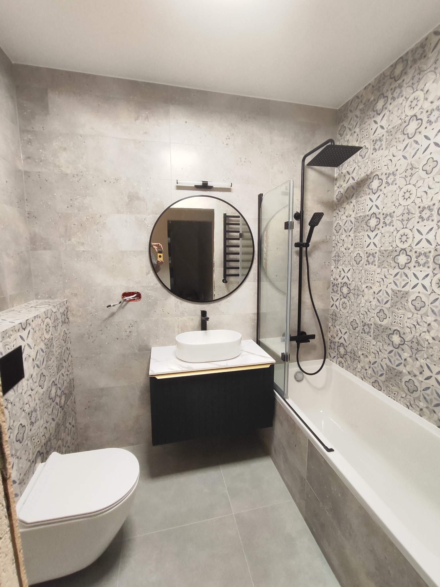 Nowoczesna łazienka z szarymi płytkami imitującymi beton, okrągłym lustrem z czarną ramą, białą umywalką nablatową na czarnej szafce, baterią w kolorze czarnym, oraz wanną z parawanem.