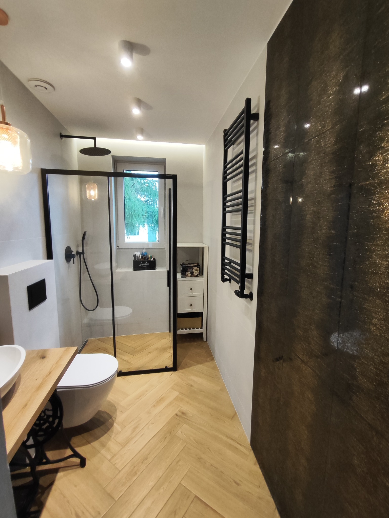 Nowoczesna łazienka z kabiną prysznicową typu walk-in z czarnymi ramami, drewnianą podłogą ułożoną w jodełkę oraz umywalką na metalowym stelażu, oświetlona punktowymi lampami sufitowymi i wiszącą...