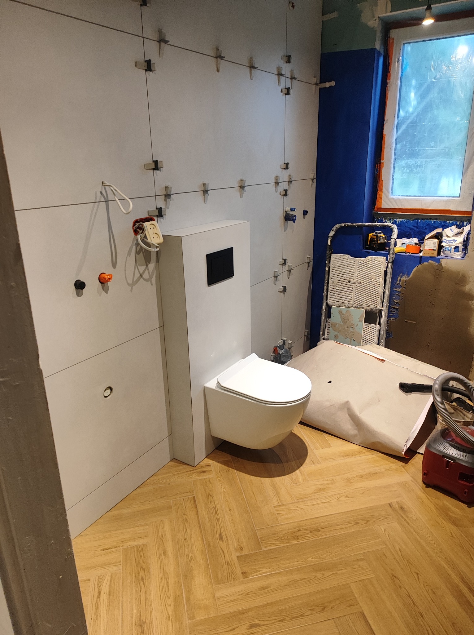 Wnętrze łazienki w trakcie prac: podwieszana toaleta, szare płytki ścienne z krzyżykami poziomującymi, drewnopodobna podłoga w jodełkę, niebieska ściana, drabina z narzędziami i materiałami...