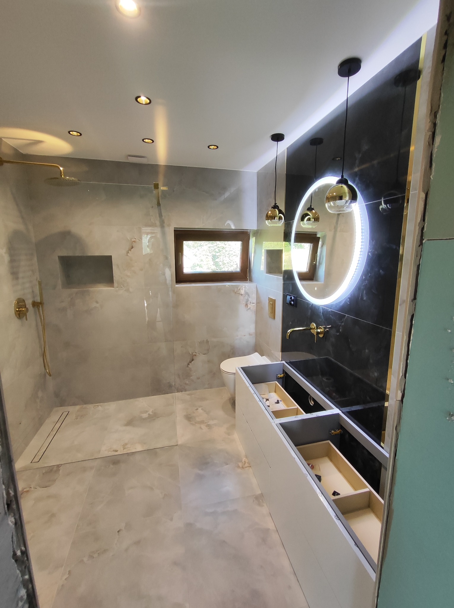 Wnętrze łazienki w trakcie prac wykończeniowych, widoczna kabina prysznicowa walk-in z deszczownicą w kolorze złotym, podświetlane lustro okrągłe, czarna ściana oraz biała szafka pod umywalkę...