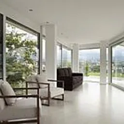 Jasne, przestronne wnętrze z dużymi przeszkleniami, widok na zieleń i miasto w tle. Dwa beżowe fotele z drewnianymi ramami i ciemna sofa tworzą minimalistyczną aranżację.