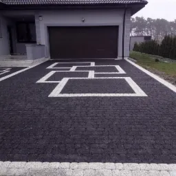 Czarna kostka brukowa z białymi, geometrycznymi wzorami na podjeździe przed nowoczesnym domem z brązową bramą garażową i zadbanym trawnikiem w tle.