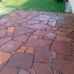 Wyłożony nieregularnymi, brązowo-czerwonymi płytami kamiennymi taras przylegający do ściany budynku z roletą zewnętrzną, widok na trawnik.