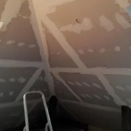 Poddasze w trakcie wykańczania: płyty gipsowo-kartonowe zamontowane na skosach dachu, widoczne łączenia wzmocnione taśmą, szpachlowane wkręty, drabina oparta o ścianę, okna dachowe po bokach.