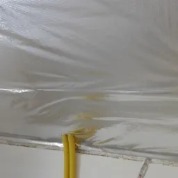 Błyszcząca folia izolacyjna na poddaszu, widoczne żółte rury i fragment drabiny, przygotowanie do ocieplenia.