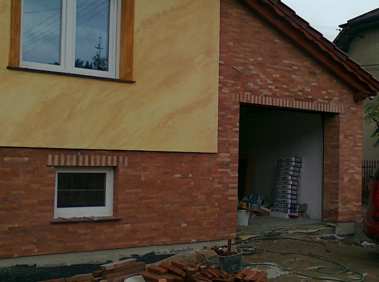 Budynek z częściowo wykończoną elewacją: tynk strukturalny w odcieniach beżu i ceglana okładzina, widoczne okna z białymi ramami i sterta worków w otwartym garażu.