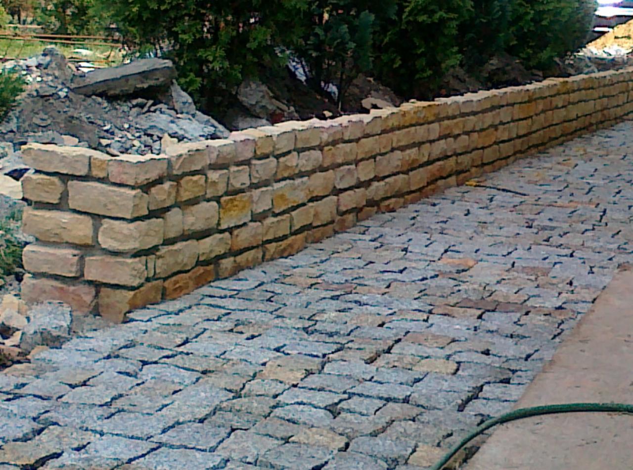 Kamienny murek oporowy z piaskowca i ścieżka z kostki brukowej, widok z perspektywy chodnika, częściowo widoczne rośliny w tle.