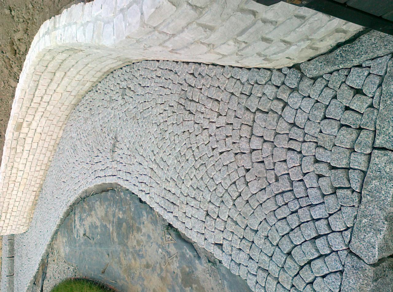 Kaskadowe ułożenie kostki granitowej w dwóch odcieniach szarości, tworzące dekoracyjną obwódkę rabaty lub ścieżki w ogrodzie.