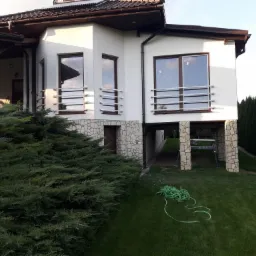 Dwukondygnacyjny dom z białymi ścianami i brązowym dachem, z balkonami zabezpieczonymi poziomymi metalowymi barierkami, otoczony zielenią, w tle rząd wysokich drzew.