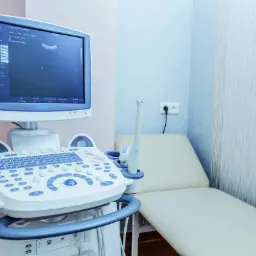 Aparat USG z widocznym ekranem prezentującym obraz, obok leżanka w jasnym gabinecie lekarskim.