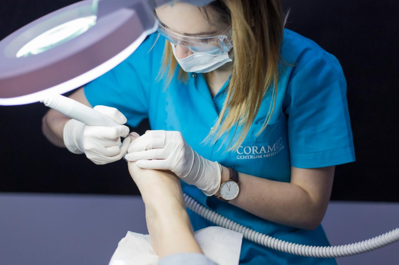 Podolog w niebieskim uniformie, masce i okularach ochronnych, precyzyjnie opracowuje paznokcie stopy klienta przy użyciu frezarki pod lampą powiększającą. Widoczny zegarek na nadgarstku podologa...