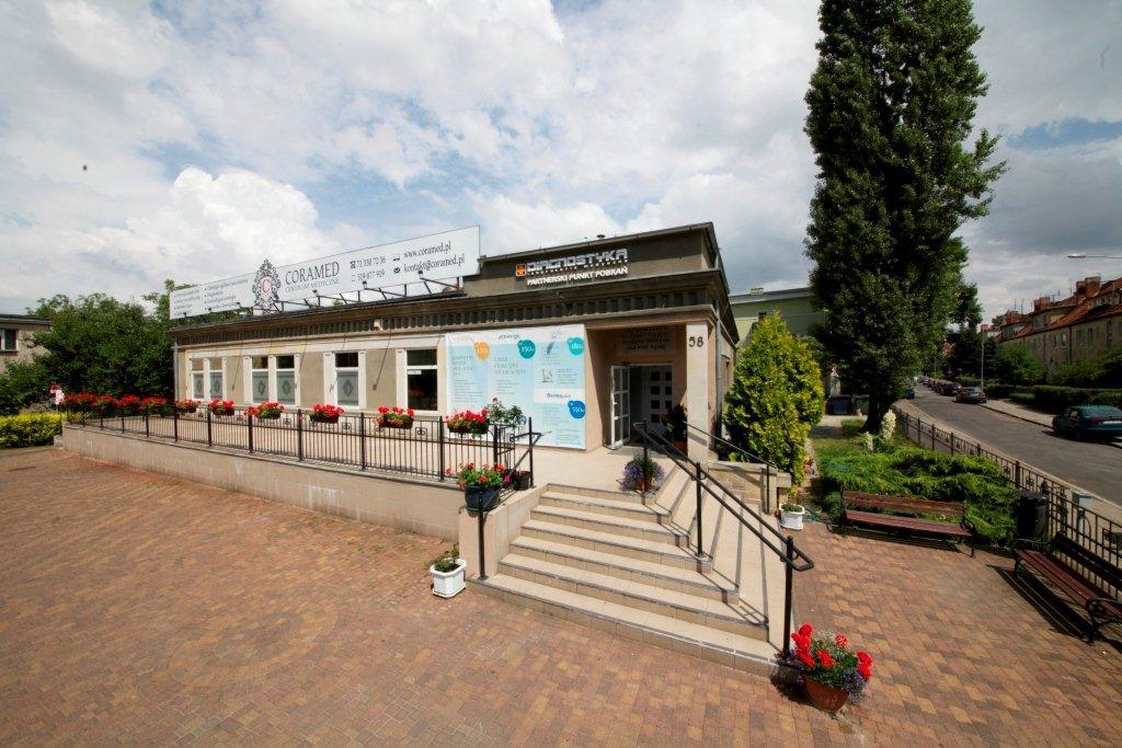 Widok z zewnątrz na budynek prywatnej kliniki z napisem 'Diagnostyka Partnerski Punkt Pobrań' nad wejściem, ozdobiony kwiatami w donicach i banerami informacyjnymi, z chodnikiem z kostki brukowej...