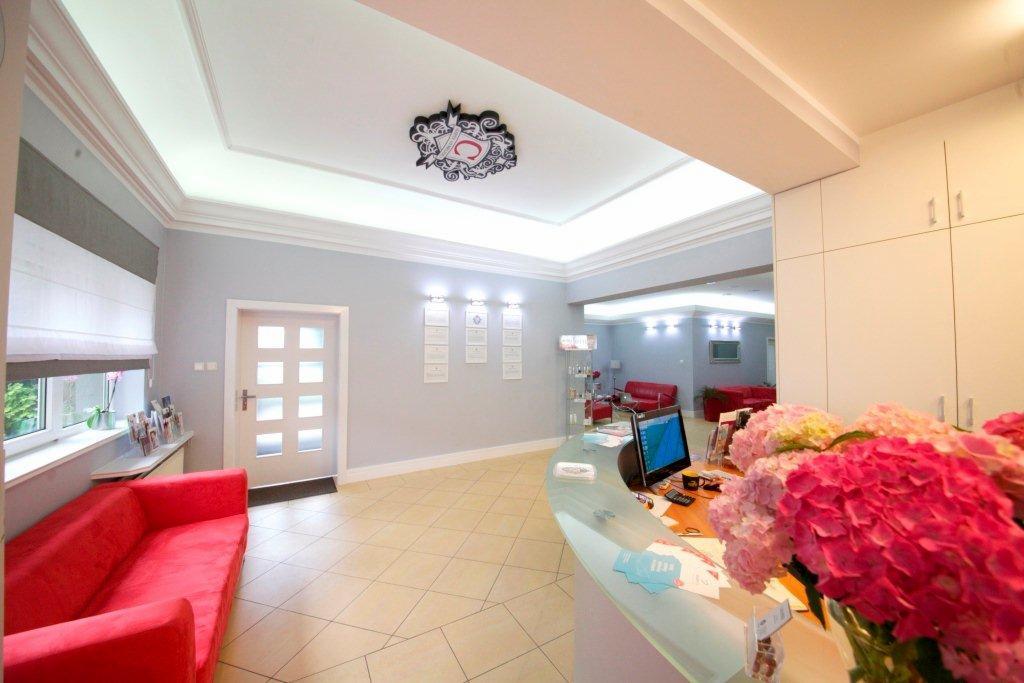 Jasne, przestronne wnętrze prywatnej kliniki z recepcją, czerwonymi sofami w poczekalni, dekoracyjnym logo na suficie i kwiatami na ladzie recepcyjnej.