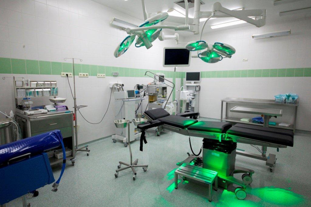 Nowoczesna sala operacyjna z zielonym oświetleniem, wyposażona w stół operacyjny, monitory, lampy zabiegowe i wózek z narzędziami medycznymi.