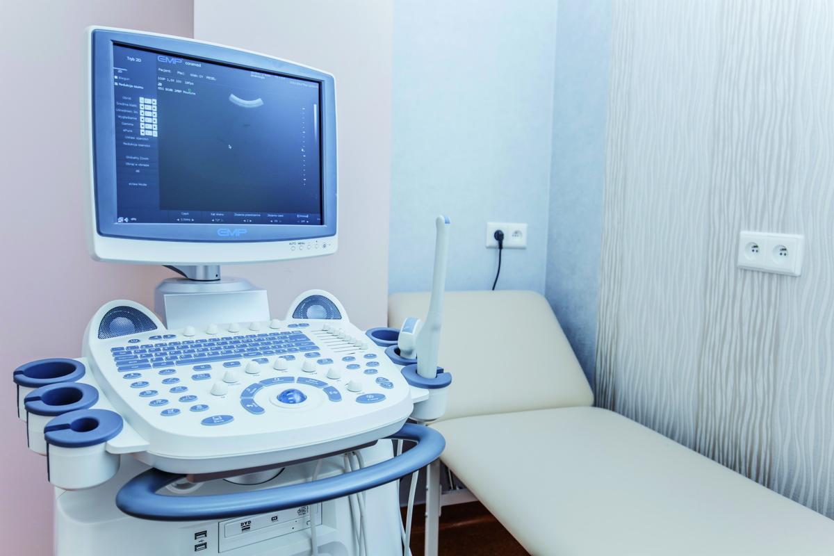 Aparat USG z widocznym ekranem prezentującym obraz, obok leżanka w jasnym gabinecie lekarskim.