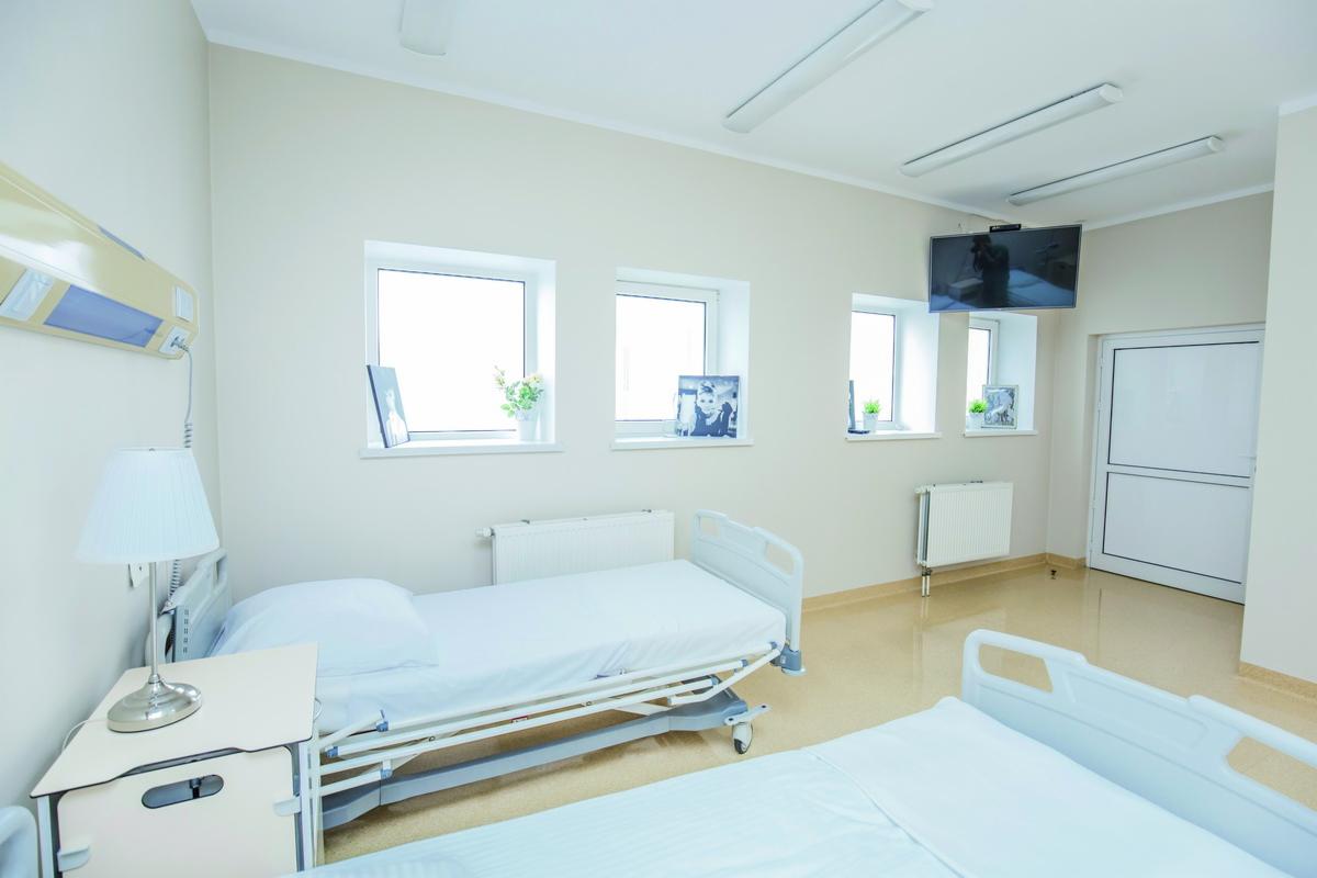 Jasne, przestronne wnętrze pokoju pacjenta w klinice we Wrocławiu, z dwoma łóżkami szpitalnymi, stolikiem nocnym z lampką, trzema oknami z dekoracjami i telewizorem na ścianie.