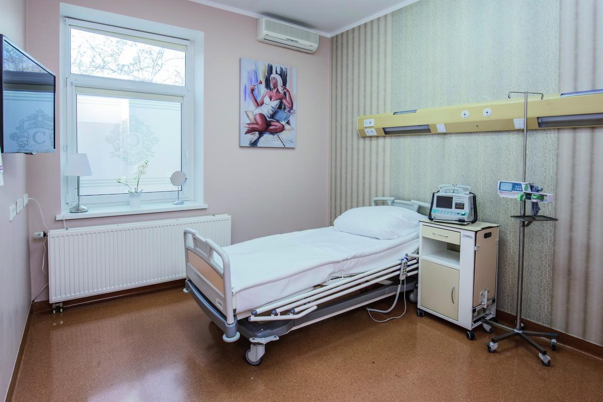 Jasne, czyste pomieszczenie szpitalne z łóżkiem regulowanym, szafką nocną z aparaturą medyczną, kroplówką, obrazem na ścianie i dużym oknem z widokiem na drzewa.