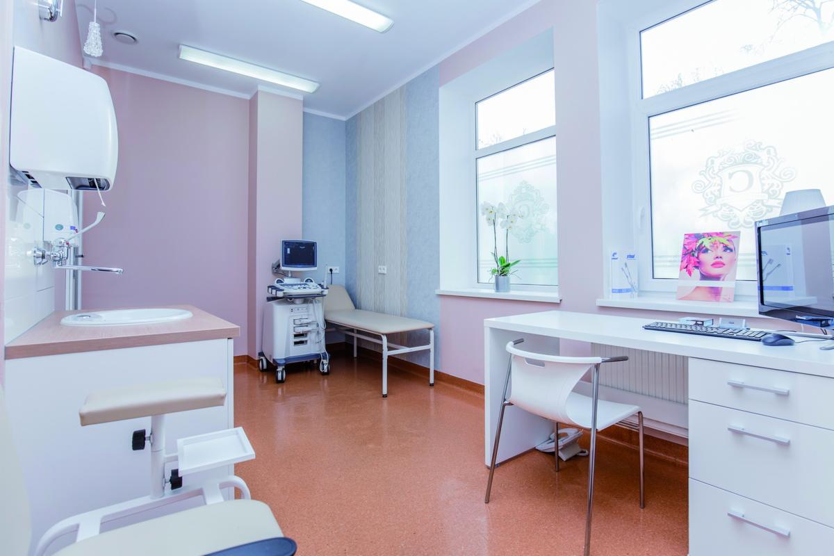 Jasne, przestronne wnętrze gabinetu lekarskiego z nowoczesnym sprzętem diagnostycznym, umywalką z fotelem zabiegowym, biurkiem z komputerem i dekoracyjnymi naklejkami na oknach.