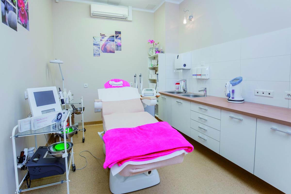 Jasne wnętrze gabinetu kosmetycznego z fotelem zabiegowym przykrytym różowym ręcznikiem, w tle białe szafki z umywalką i sprzęt kosmetyczny na wózku.