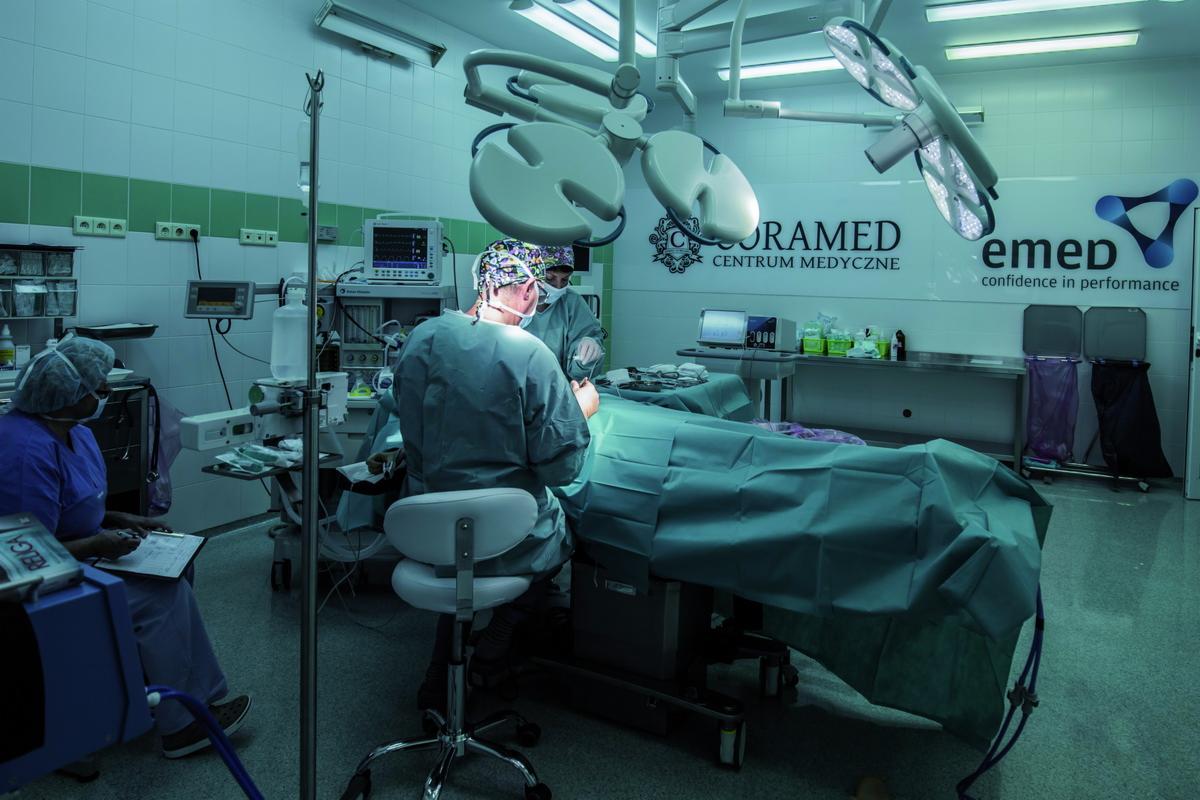 Sala operacyjna z dwoma chirurgami w trakcie zabiegu, widoczne monitory medyczne, lampy operacyjne i instrumenty, w tle logo centrum medycznego.