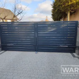 Nowoczesna, przesuwna brama z szarych paneli, z widocznym mechanizmem automatyki, zamontowana na posesji z kostki brukowej.