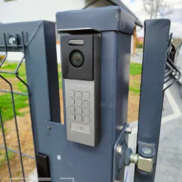 Szary wideodomofon Vidos z klawiaturą numeryczną zamontowany na metalowym słupku bramy posesji.