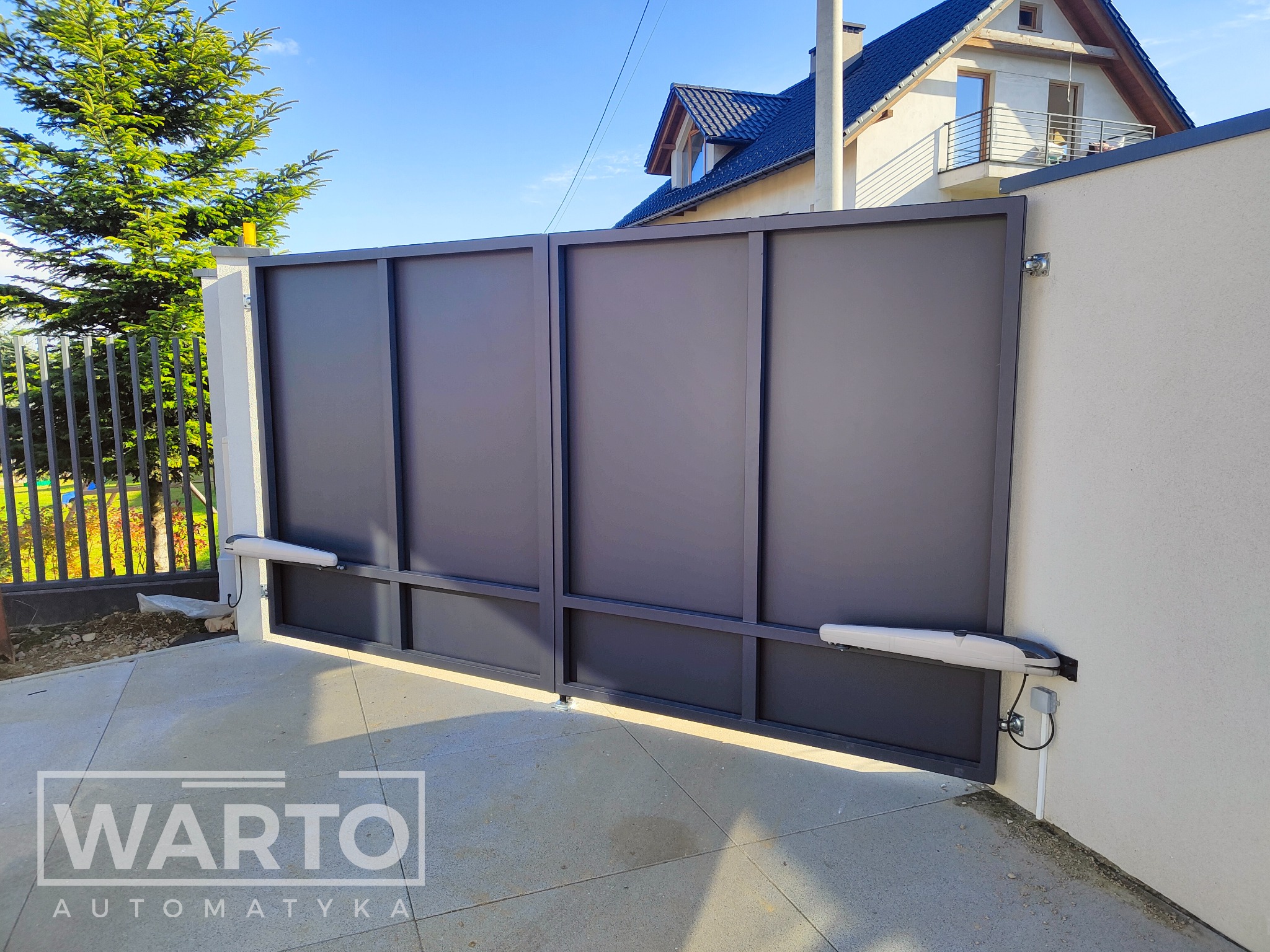 Nowoczesna, dwuskrzydłowa brama w kolorze grafitowym z zamontowaną automatyką, widok z zewnątrz na tle jasnego domu z balkonem i fragmentem zielonego drzewa.