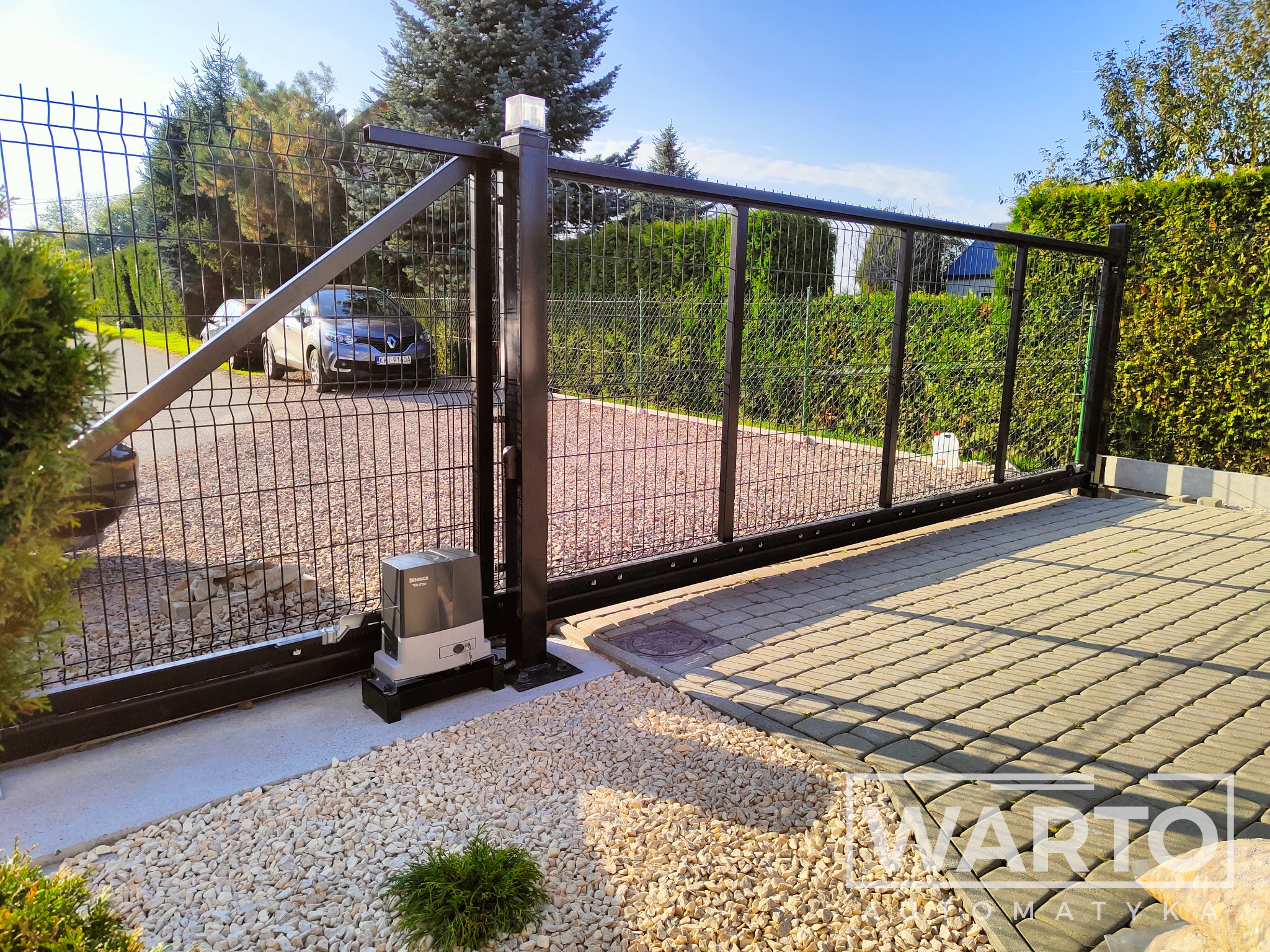 Zewnętrzna, przesuwna brama wjazdowa z czarnej siatki, zautomatyzowana napędem Beninca TO.GO, widok na podjazd z kostki brukowej i samochód za bramą.