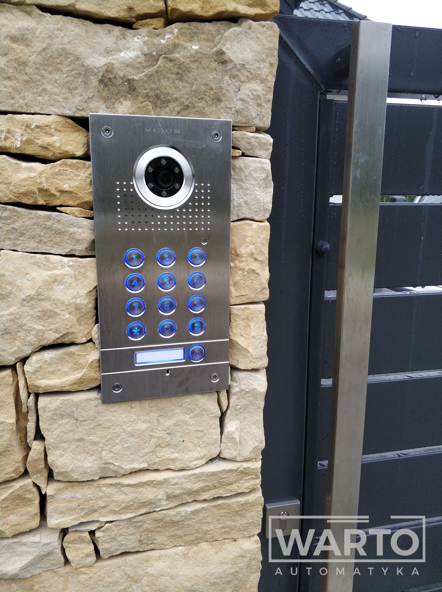 Zewnętrzny panel wideodomofonu Vidos zamontowany na kamiennym ogrodzeniu obok nowoczesnej, grafitowej bramy wjazdowej z elementami ze stali nierdzewnej.
