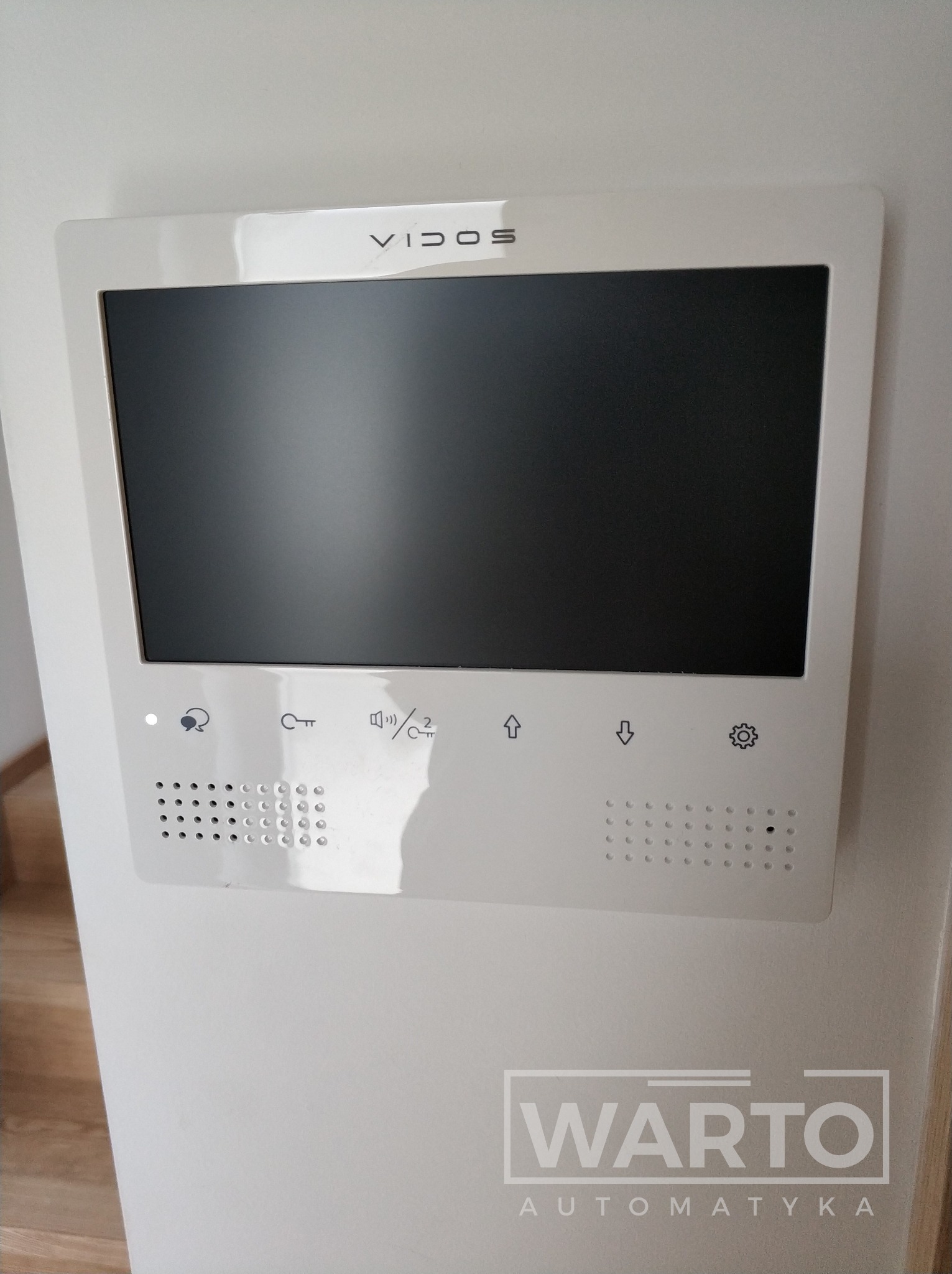 Biały wideodomofon marki Vidos zamontowany na ścianie, z widocznym ekranem i przyciskami funkcyjnymi. W dolnej części logo firmy WARTO Automatyka.