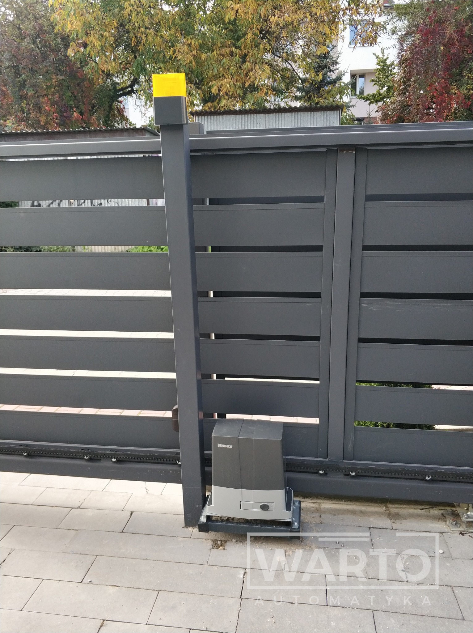 Szara, przesuwna brama automatyczna z napędem Beninca i żółtą lampą ostrzegawczą, widoczna na tle jesiennego ogrodu.
