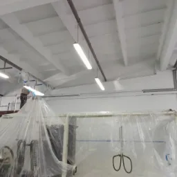 Pracownik na rusztowaniu, w trakcie prac wykończeniowych wnętrza hali, zabezpieczone folią maszyny, sufit z widocznymi belkami i oświetleniem.