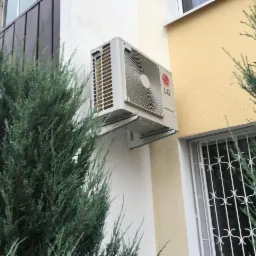 Zewnętrzna jednostka klimatyzacji LG zamontowana na ścianie budynku, widoczna zza gęstych krzewów iglastych.