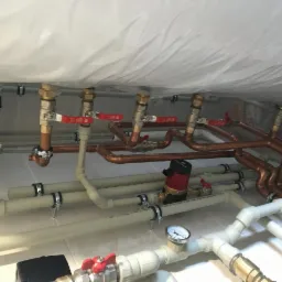 Kompleksowa instalacja hydrauliczna z rur miedzianych i PCV, widoczne zawory z czerwonymi uchwytami, manometr i naczynie przeponowe, całość pod stropem zabezpieczonym folią.