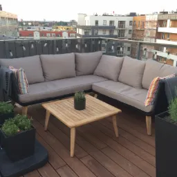 Kącik wypoczynkowy na tarasie z narożną sofą w kolorze beżowym, drewnianym stolikiem kawowym i roślinami w czarnych donicach, oświetlony girlandą żarówek, z widokiem na zabudowę miejską w tle.