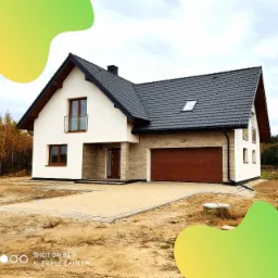 Nowoczesny dom jednorodzinny z garażem w Piskórce, wykończony jasnym tynkiem i elementami z kamienia dekoracyjnego, z ciemną dachówką i drewnianą bramą garażową.