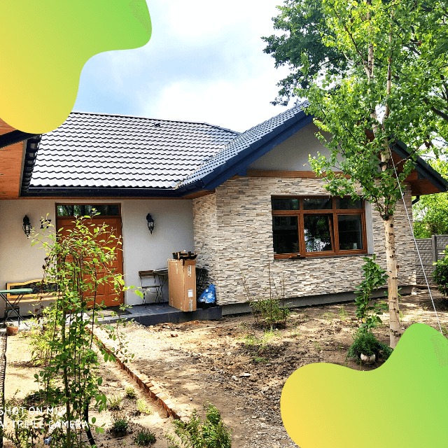 Nowoczesny dom jednorodzinny z ciemną dachówką, elewacją z kamienia dekoracyjnego i drewnianą stolarką okienną, otoczony młodymi nasadzeniami w surowym ogrodzie.