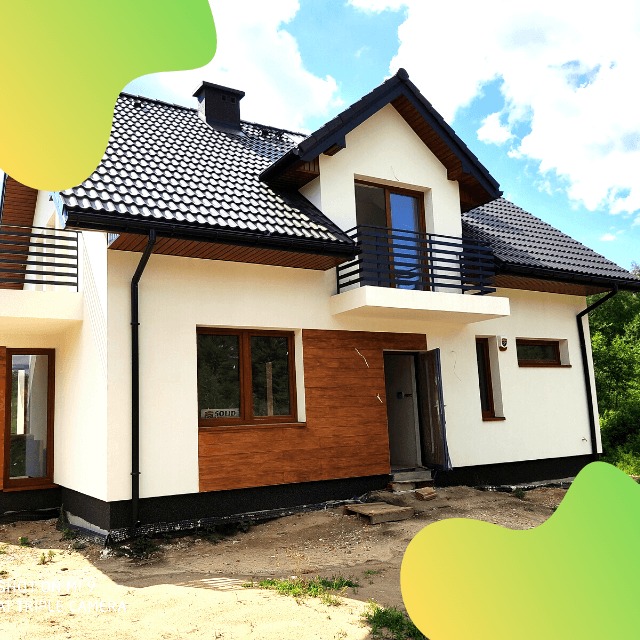 Dwupiętrowy dom murowany z czarną dachówką, drewnianą okładziną na elewacji i balkonem z czarną balustradą, w trakcie prac wykończeniowych wokół budynku.