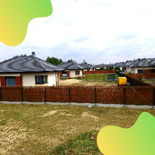 Osiedle nowych domów jednorodzinnych z ciemnymi dachami, ogrodzone niskim, brązowym płotem z wikliny, widok z trawnika w pochmurny dzień.