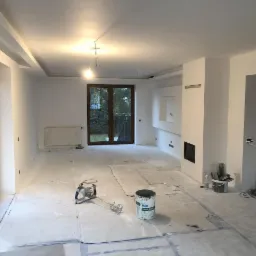 Przestronne wnętrze w trakcie prac wykończeniowych: zabezpieczona podłoga, ściany przygotowane do malowania, widoczny agregat do gładzi i wiadro z masą szpachlową Knauf Super Finish.