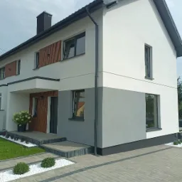 Dwukondygnacyjny dom z elewacją w odcieniach szarości i bieli, z drewnianymi akcentami i nowoczesną kostką brukową na podjeździe.
