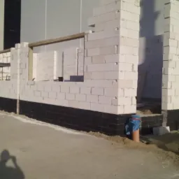 Wznoszenie ścian z białych bloczków na czarnej hydroizolacji fundamentów w Zalewie, widoczne otwory okienne i drzwiowe w surowym murze.