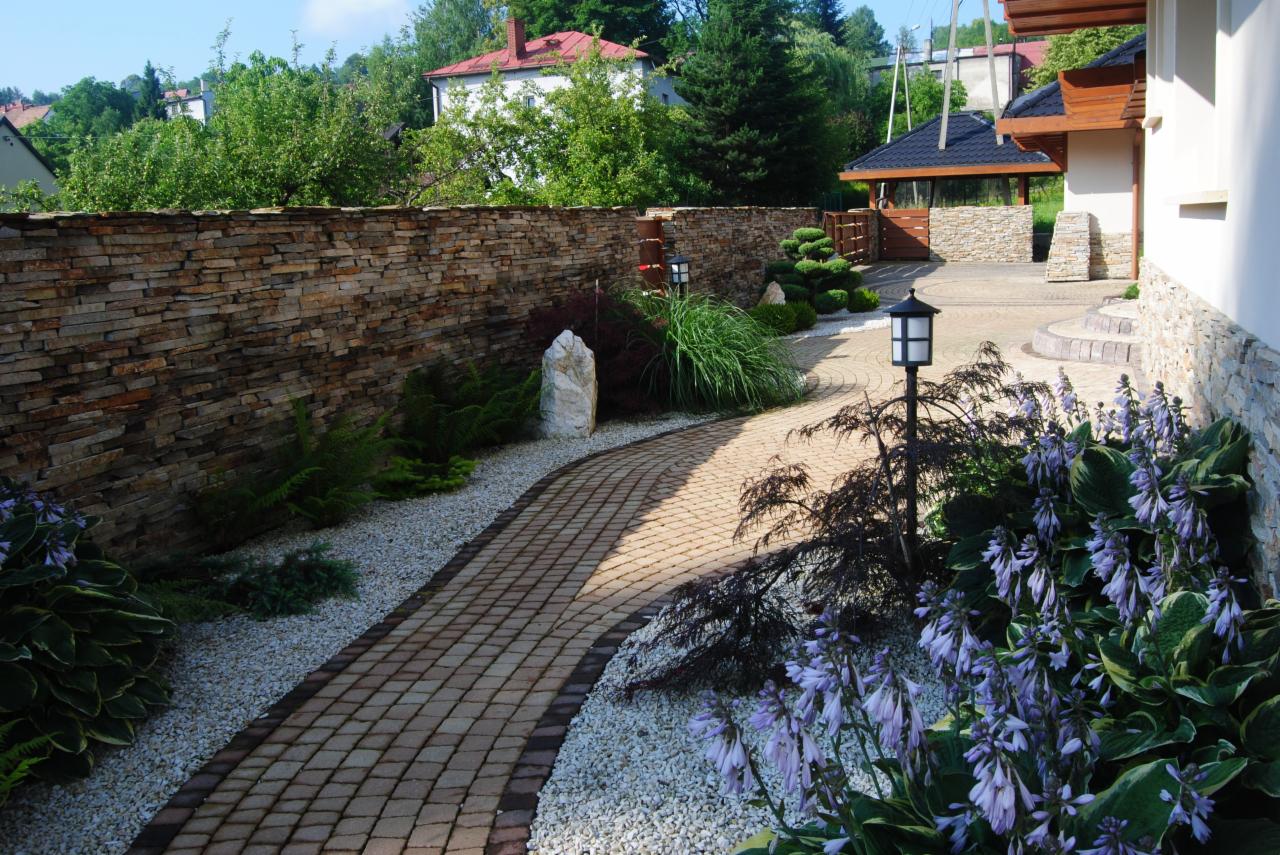 Ogród z brukowaną ścieżką wzdłuż kamiennego muru, ozdobiony białym żwirem, lampami ogrodowymi i kwitnącymi funkiami.